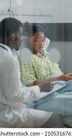 Young Patient Receiving Medical Consultation From Doctor In Hospital Ward. Medical Specialist Talking To Ill Woman In Bed About Prescription Treatment And Healthcare To Heal Disease.