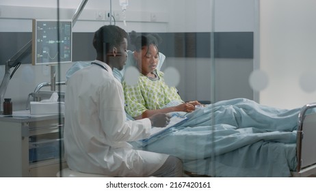 Young Patient Receiving Medical Consultation From Doctor In Hospital Ward. Medical Specialist Talking To Ill Woman In Bed About Prescription Treatment And Healthcare To Heal Disease.