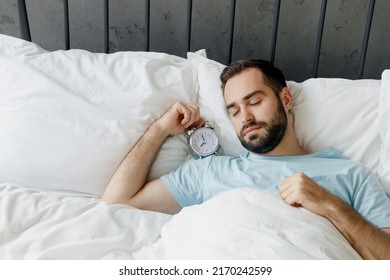 Young Peaceful Calm Sleepy Unshaven Man Wear Casual Blue T-shirt Lying In Bed Sleep Hold Clock Alarm Rest Relax Spend Time In Bedroom Lounge Home In Own Room House Wake Up Dream. Real Estate Concept