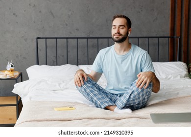Young peaceful man in casual blue t-shirt sitting on bed in lotus pose yoga om aum gesture relax meditate try to calm down listen music in earphones rest relax spend time in bedroom home in own room