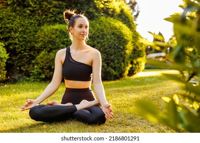 Young Peaceful Woman Sitting In Lotus Pose, Breathing And Meditating. Concept Of Outdoors Yoga, Relaxation, Zen And Mental Balance. Beautiful Yogini In Black Sportswear In The Park At Sunset.