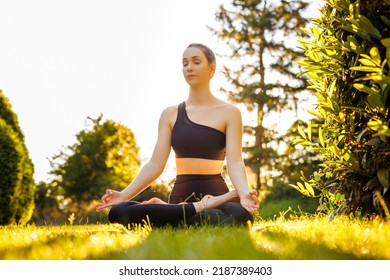 Young Peaceful Woman Sitting In Lotus Pose, Breathing And Meditating. Concept Of Outdoors Yoga, Relaxation, Zen And Mental Balance. Beautiful Yogini In Black Sportswear In The Park At Sunset.