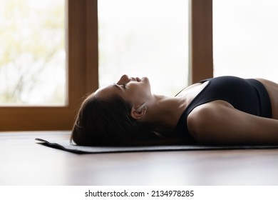 Young peaceful woman wearing black sports top practicing yoga lying in Dead Body pose on mat, doing Corpse Savasana exercise, relaxing after workout at home, meditating listen music through earbuds - Powered by Shutterstock