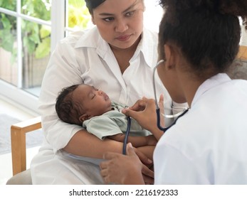 Young Pediatrician Checking The Well Being Of Little Baby Boy Who's Sleeping In Mother's Arms. Healthcare And Well Being Concept.