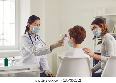 Young Pediatrician In Medical Face Mask Measuring Little Boy's Body Temperature With Non-contact Infrared Electronic Thermometer. Mother Calming Down Her Child During Checkup At Family Doctor's Office