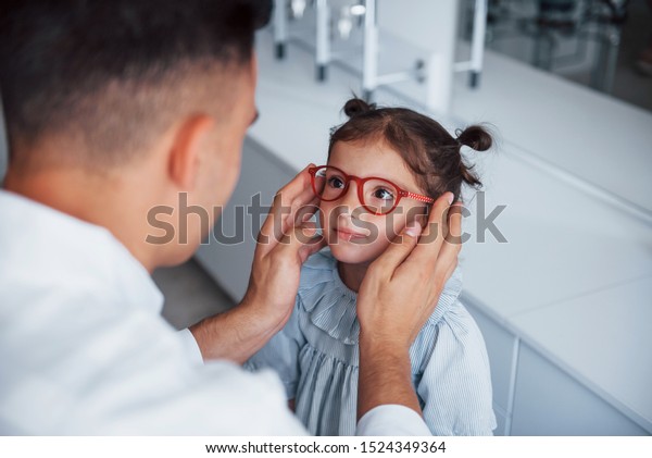 白いコートを着た小児科医は、小さな女の子に新しい眼鏡を買うのに役立ちます。