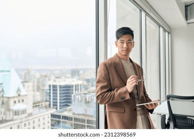 Young pensive Asian business man professional manager executive leader holding digital tablet standing in corporate office looking at window with city view, thinking of solution, lost in thoughts. - Powered by Shutterstock