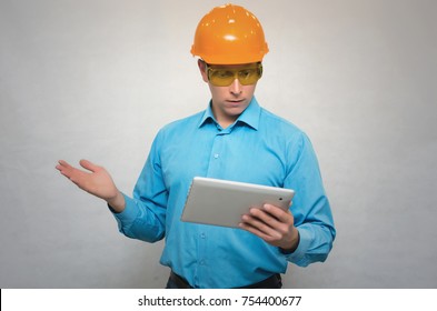 Young Pensive Builder Worker In A Hard Hat Holding In Hand A Tablet Computer Looking On It And Thinking Isolated. Engineer Learning Construction Plans. The Obvious Solution.