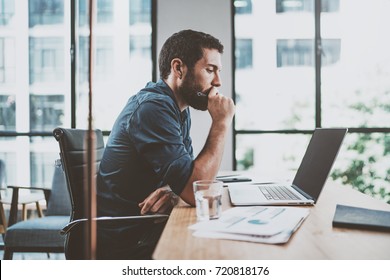 Young pensive coworker working at sunny work place loft while sitting at the wooden table.Man analyze document on laptop display.Blurred background.Horizontal