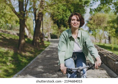 Young pensive dreamful happy woman 20s wearing casual green jacket jeans riding bicycle bike on sidewalk in city spring park outdoors, look aside. People active urban healthy lifestyle cycling concept