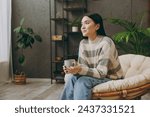 Young pensive happy woman wears casual clothes sits on armchair hold cup drink coffee look camera stay at home hotel flat rest relax spend free spare time in grey living room indoor. Lounge concept