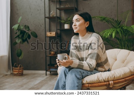 Young pensive happy woman wears casual clothes sits on armchair hold cup drink coffee look camera stay at home hotel flat rest relax spend free spare time in grey living room indoor. Lounge concept