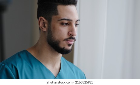 Young Pensive Hispanic Doctor Male Thoughtful Arabian Nurse Practitioner Therapist Surgeon Stand Near Window In Hospital Workplace Wait For Patient Visit Sad Medical Man Think About Ending Quarantine