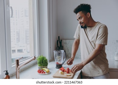 Young pensive man of African American ethnicity wear casual clothes prepare sweet breakfast cut fruit talk on mobile cell phone cook food in indoor kitchen at home alone Healthy diet lifestyle concept - Shutterstock ID 2211467363