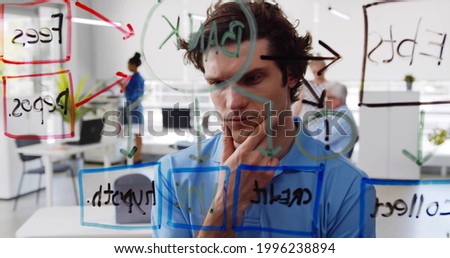 Young pensive man looking at data on glass board in modern office. Portrait of businessman brainstorming reading charts on glassboard in creative coworking workspace
