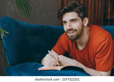 Young Pensive Man Wear Red T-shirt Writing Memories In Notebook Diary Lay Down On Blue Sofa Stay Home Hotel Flat Rest Relax Spend Free Spare Time In Living Room Indoor Grey Wall People Lounge Concept