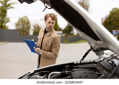 Young pensive mechanic making diagnostic, standing near car with open hood. Repair, car service concept  - Shutterstock ID 2256853437