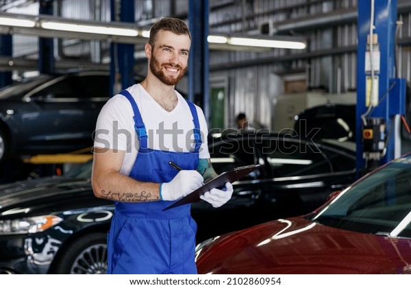 Young pensive professional car mechanic man in denim\
blue overalls white t-shirt gloves hold clipboard with papers\
document work in modern vehicle repair shop workshop indoors.\
Tattoo translate fun.