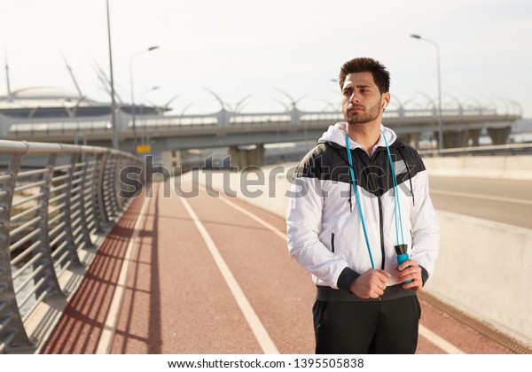 Young pensive\
sportsman in windbreaker and sports pants looking at riverside\
while having break after\
training