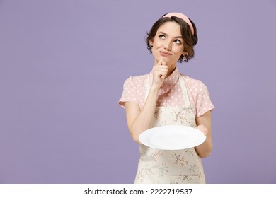 Joven pensante ama de casa ama de casa cocinera cocinera mujer panadera en delantal rosa sosteniendo un plato blanco vacío apuntando a la barbilla se ve aislado en un fondo violeta pastel Concepto de proceso de cocina