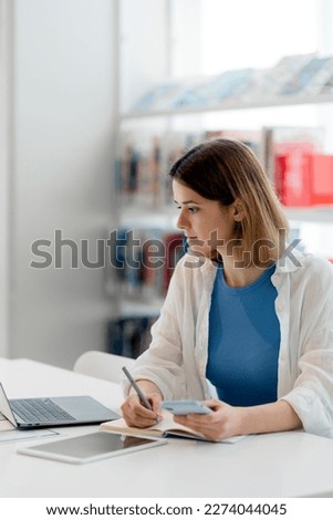 Young pensive university student studying, taking notes using laptop computer and mobile phone sitting in library. Online education concept