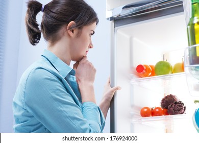 Young Pensive Woman Looking In The Refrigerator With Hand On Chin.