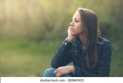 Young pensive woman relaxing at the park and sitting on grass, vegetation on background