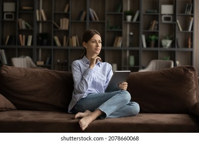 Young Pensive Woman Sit On Sofa With Digital Tablet, Buying On E-commerce Retail Services Consider Purchase, Thinks Over Commercial Offer Spend Weekend Alone At Home. Modern Tech, Lifestyle Concept