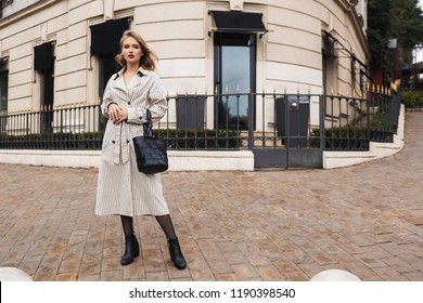 Young pensive woman in striped trench coat with black handbag thoughtfully looking in camera walking around cozy city street - Shutterstock ID 1190398540