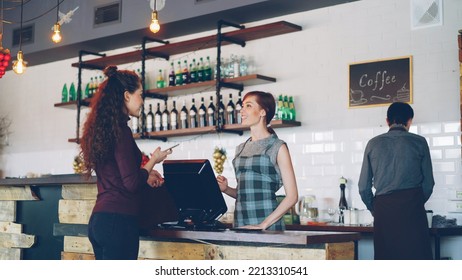 Young people are buying coffee-to-go in nice local cafe and paying with smartphone while friendly workers are greeting customers, talking and selling drinks.