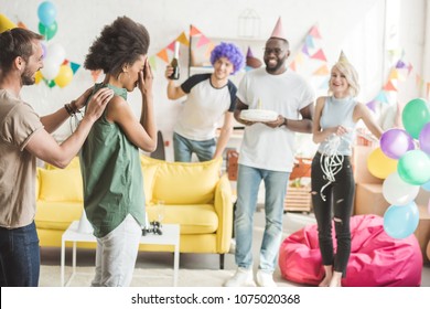 Young People Covering Eyes Of Young Woman And Greeting Her With Birthday Cake  On Surprise Party