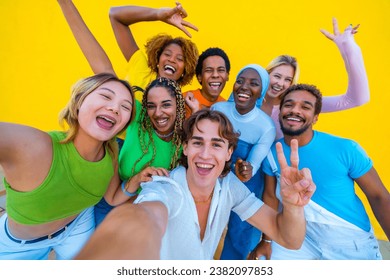 Young people from different ethnicities taking a happy selfie at a gathering next to a yellow background - Powered by Shutterstock