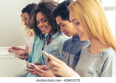 Young People Of Different Nationalities Using Smartphones And Smiling While Sitting In A Row. Beautiful Afro American Girl Is Looking At Camera