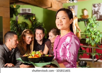 Young people eating in a Thai restaurant; the waitress brings the dishes, rice and others