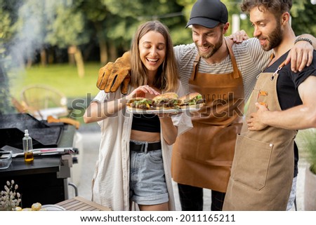 Young people enjoy yummy burgers made on a grill at picnic, standing together and having fun. Friends cooking at backyard outdoors. American lifestyle