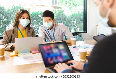 Young People With Face Masks Back At Work In Office After Lockdown.Asian Small Business Startup Multiracial Brainstorm Meeting With Laptop And Chart Paper Everyone Mask For Covid19 Protection Corona.