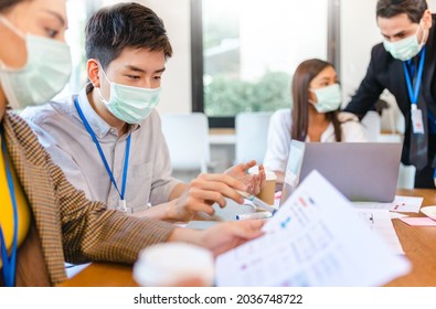 Young People With Face Masks Back At Work In Office After Lockdown.Asian Small Business Startup Multiracial Brainstorm Meeting With Laptop And Chart Paper Everyone Mask For Covid19 Protection Corona.