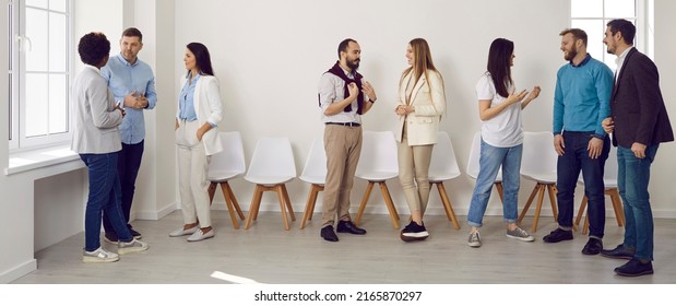 Young People Gather For A Business Meeting In The Office. Diverse Team Of Men And Women Standing And Talking In Pairs Or Groups Of Three At A Modern Business Training Event