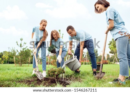 Young people girls and boy volunteers outdoors helping nature planting trees together pouring water from bucket on ground smiling cheerful