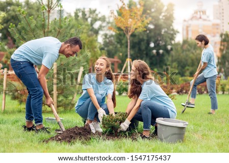 Young people girls and boy volunteers outdoors helping nature planting trees digging ground with shovel talking smiling cheerful