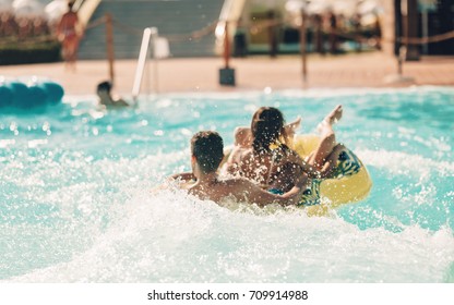 Young people having fun on the water slide with friends and familiy in the aqua fun park glides, happy falling into water and water splashes are all over. Blue sky background looks amazing sunlight