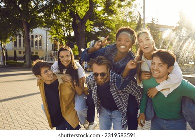 Young people having fun together. Group of six positive joyful diverse mixed race friends meet on good sunny day in city. Happy men piggyback beautiful girls who are posing for photo and doing V signs