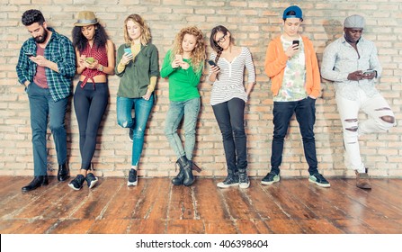 Young People Looking Down At Cellular Phone - Teenagers Leaning On A Wall And Texting With Their Smartphones - Concepts About Technology And Global Communication