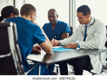 Young people on medical staff meeting, with man in white coat showing something in journal, sitting at table in bright room - Powered by Shutterstock