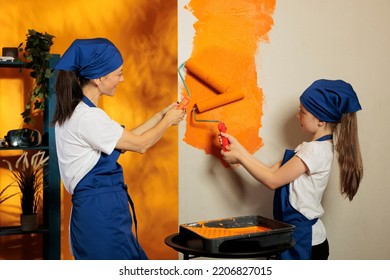 Young People Painting Orange Walls At Home, Using Color Paint And Roller Paintbrush To Work On Apartment Renovation. Woman With Little Child Redecorating House Interior With Tools.