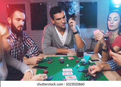 Young people play poker at the table. On the table they have glasses with alcoholic beverages, mobile phones and chips for the game. One of the players is smoking a cigar.
