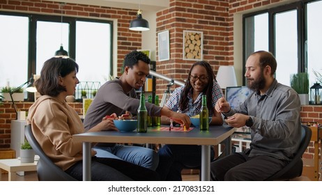 Young People Playing Strategic Board Games With Cards, Rolling Dice To Move Figurines. Men And Women Gathering To Have Fun And Play Charades Game In Living Room, Having Beer Bottles And Snacks.