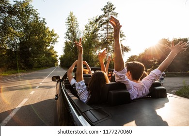 Сompany of young people riding in a cabriolet on the road and holding their hands up on a warm sunny day. Back view. - Powered by Shutterstock