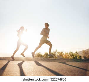 Young People Running On Country Road With Bright Sunlight. Outdoor Shot Of Young Man And Woman Jogging In Morning.