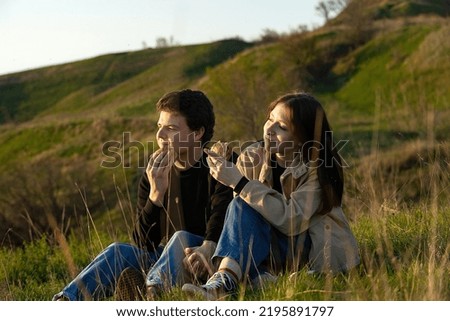 young people students, a guy and a girl in love are sitting in nature at sunset outside the city talking and eating hamburgers